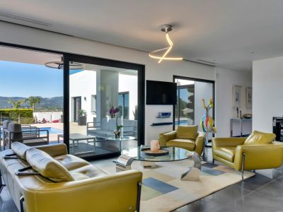 Luminoso salón con sofá y sillones de cuero verde oliva, mesa de centro de cristal y acceso directo a la terraza, piscina y vistas a la montaña.