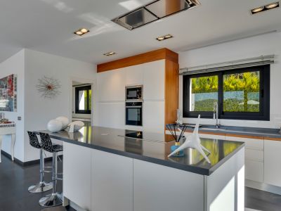 Moderna cocina con gran isla central de encimera oscura y placa integrada, armarios blancos sin tiradores y electrodomésticos empotrados. Ventana amplia con vistas a la vegetación.