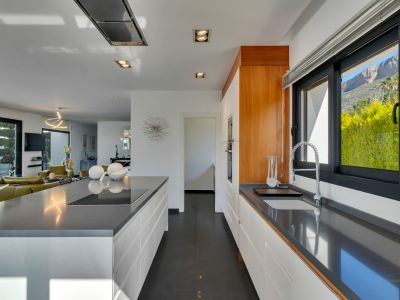 Moderna cocina de planta abierta con encimeras de cuarzo gris, armarios blancos sin tiradores, placa de inducción integrada en la isla y vistas a la montaña desde la ventana.