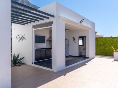 Moderna cocina exterior y zona de barbacoa con mobiliario oscuro, integrada con un práctico baño exterior, todo en un patio embaldosado con vistas a la montaña.