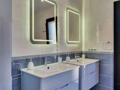 Moderno baño con doble lavabo suspendido de color azul claro, encimeras blancas y dos espejos retroiluminados LED sobre paredes alicatadas en tonos azules y grises.