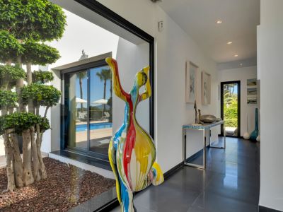 Moderno pasillo con suelo oscuro, escultura abstracta colorida y vistas a la terraza con piscina y un árbol ornamental interior.