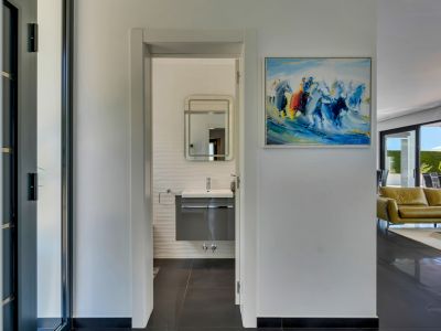 Pasillo luminoso que conduce a un baño moderno con tocador flotante, paredes texturizadas y espejo iluminado. Un cuadro abstracto adorna la pared, con vistas parciales al salón y terraza.
