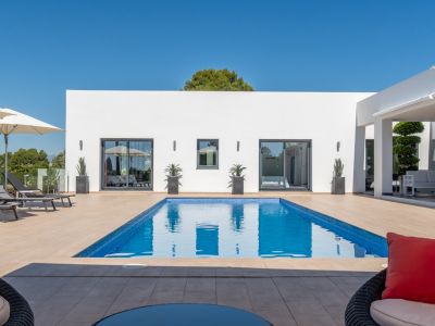 Piscina rectangular de mosaico azul rodeada de una terraza de gres, frente a una moderna fachada blanca con amplias puertas correderas y un salón exterior cubierto.
