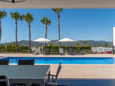 Terraza cubierta con mesa de comedor exterior, con vistas a la piscina de azulejos azules y al paisaje montañoso con palmeras bajo un cielo despejado.