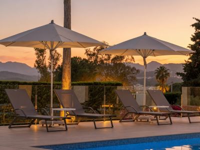 Terraza junto a la piscina con cuatro tumbonas y dos sombrillas iluminadas al atardecer, ofreciendo vistas a las montañas y un ambiente sereno.