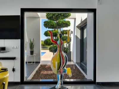 Una escultura moderna y colorida enmarca la vista desde el salón a través de un gran ventanal negro hacia un jardín cuidado con un árbol escalonado y piscina.