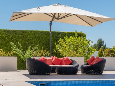 Zona de estar exterior con mobiliario de ratán sintético negro y cojines rojos, bajo una sombrilla, junto a la piscina de mosaico azul.