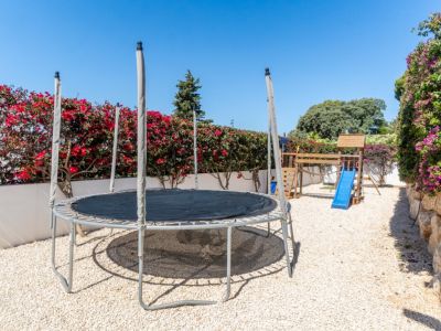 Zona de juegos exterior con cama elástica y estructura de madera con tobogán azul sobre gravilla. Arbustos de buganvilla y un muro de piedra con enredaderas moradas enmarcan el espacio bajo un cielo despejado.