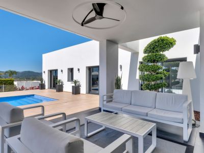 Zona lounge exterior cubierta con mobiliario blanco moderno, ventilador de techo y acceso directo a la piscina. Arquitectura minimalista y vistas a la montaña.