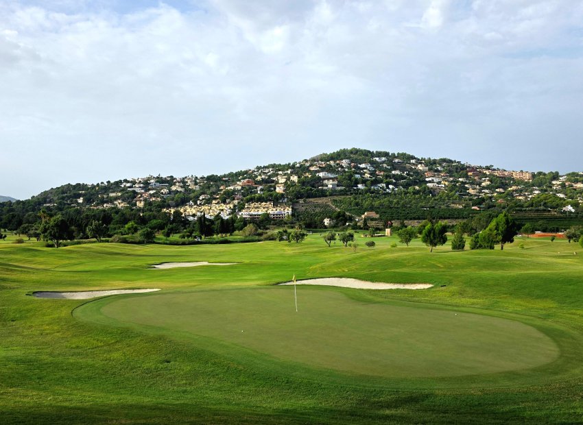 Amplio green de campo de golf con búnkeres de arena y bandera, enmarcado por una ladera verde salpicada de residencias de lujo bajo un cielo nublado.