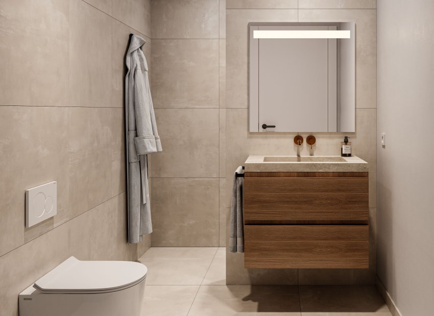 Baño moderno con mueble de lavabo flotante de madera, lavabo integrado de piedra, grifos de bronce empotrados y espejo iluminado. Azulejos beige de gran formato revisten paredes y suelo, junto a inodoro suspendido.