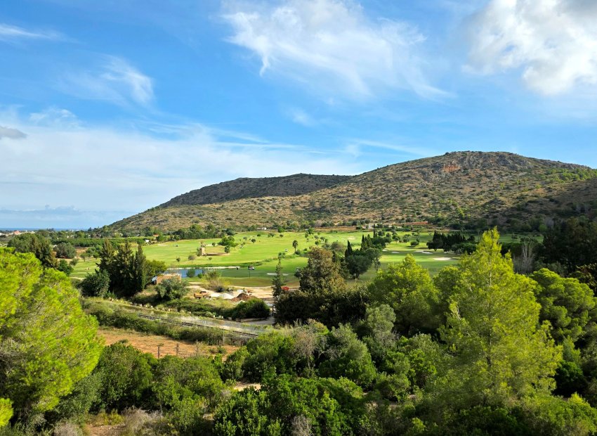 Vista panorámica de un campo de golf verde con obstáculo de agua y una torre de piedra distintiva, enmarcado por árboles, una montaña árida y el mar lejano.