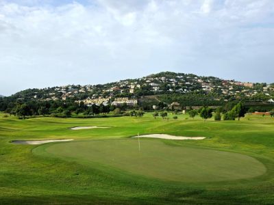 Amplio green de campo de golf con búnkeres de arena y bandera, enmarcado por una ladera verde salpicada de residencias de lujo bajo un cielo nublado.