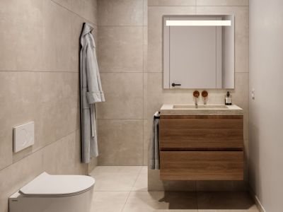 Baño moderno con mueble de lavabo flotante de madera, lavabo integrado de piedra, grifos de bronce empotrados y espejo iluminado. Azulejos beige de gran formato revisten paredes y suelo, junto a inodoro suspendido.