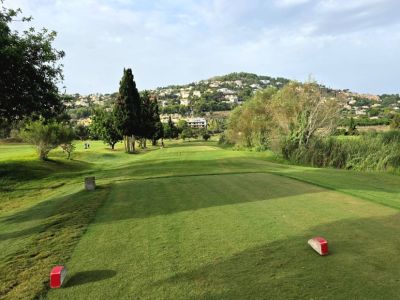 Un tee de golf impecablemente cuidado se extiende hacia un fairway verde, enmarcado por cipreses y casas en la ladera. Marcadores rojos señalan el inicio del juego en este pintoresco hoyo.
