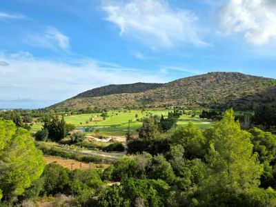 Vista panorámica de un campo de golf verde con obstáculo de agua y una torre de piedra distintiva, enmarcado por árboles, una montaña árida y el mar lejano.