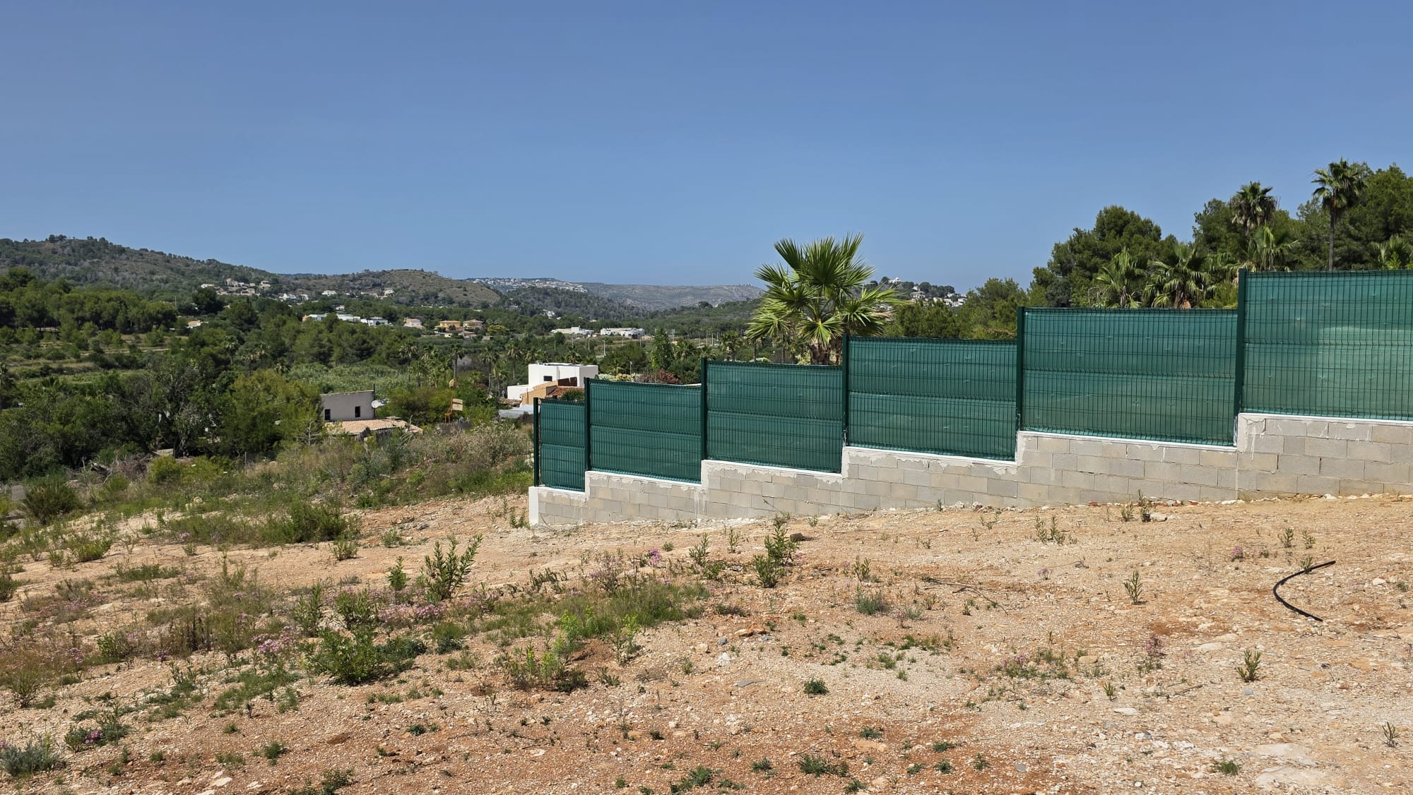 Parcela árida con valla escalonada de bloques de hormigón y malla verde. Vistas amplias hacia colinas mediterráneas con viviendas dispersas bajo un cielo despejado.