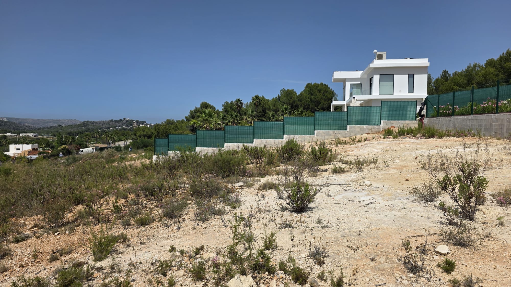 Parcela sin desarrollar con terreno seco y rocoso, vegetación escasa, junto a una residencia moderna blanca. Ofrece vistas claras hacia colinas verdes bajo un cielo azul.