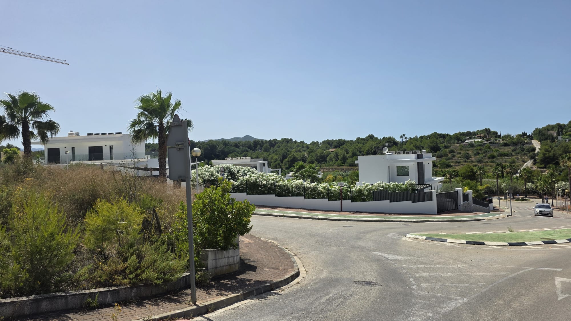 Vista de calle residencial con villas blancas modernas y balcones de cristal, rodeadas de vegetación mediterránea y colinas verdes. Infraestructura vial y cielo despejado son visibles.