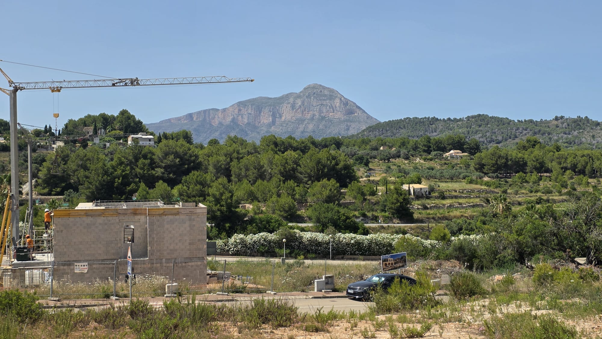 Vista de una obra con estructura de bloques de hormigón y grúa, enmarcada por la vegetación mediterránea, colinas aterrazadas y la imponente montaña del Montgó bajo un cielo azul claro.