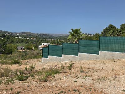 Parcela árida con valla escalonada de bloques de hormigón y malla verde. Vistas amplias hacia colinas mediterráneas con viviendas dispersas bajo un cielo despejado.