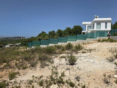 Parcela sin desarrollar con terreno seco y rocoso, vegetación escasa, junto a una residencia moderna blanca. Ofrece vistas claras hacia colinas verdes bajo un cielo azul.