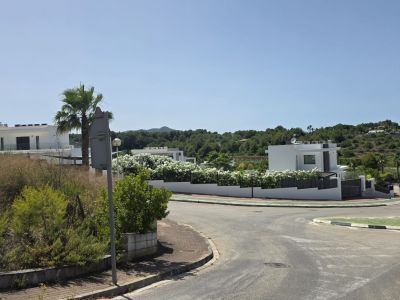 Vista de calle residencial con villas blancas modernas y balcones de cristal, rodeadas de vegetación mediterránea y colinas verdes. Infraestructura vial y cielo despejado son visibles.
