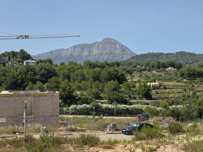 Vista de una obra con estructura de bloques de hormigón y grúa, enmarcada por la vegetación mediterránea, colinas aterrazadas y la imponente montaña del Montgó bajo un cielo azul claro.
