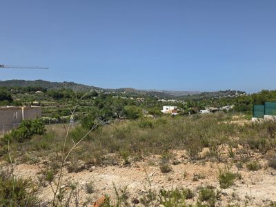 Vista de una parcela con vegetación natural y suelo árido, enmarcada por colinas verdes distantes y edificios residenciales, con una grúa de construcción visible.