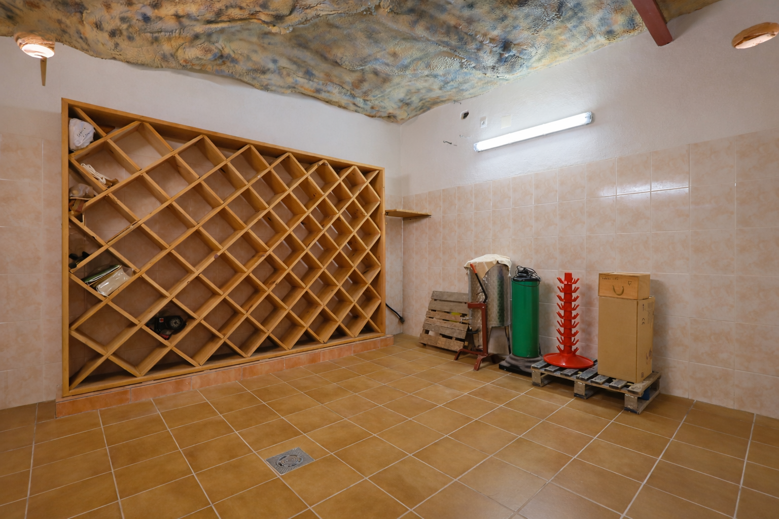 Amplia bodega con un gran botellero de madera en cuadrícula, paredes alicatadas y suelo cerámico. Destaca el techo con textura de cueva y la iluminación funcional. Amplia bodega con un gran botellero de madera en cuadrícula, paredes alicatadas y suelo cerámico. Destaca el techo con textura de cueva y la iluminación funcional.