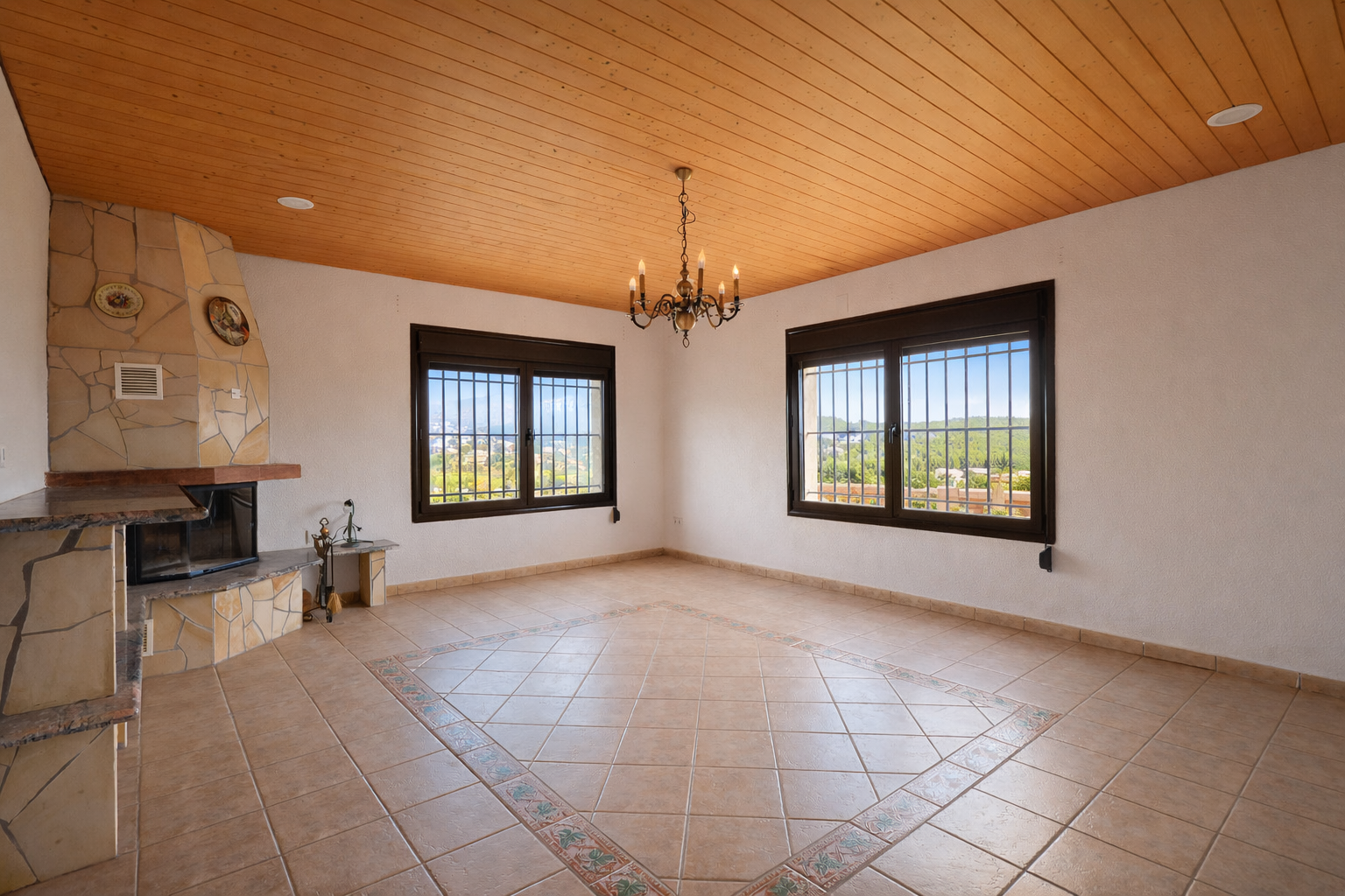 Amplia estancia con chimenea de piedra, techo de madera machihembrada y suelo de baldosas con patrón central decorativo. Dos ventanas enrejadas ofrecen vistas al valle. Amplia estancia con chimenea de piedra, techo de madera machihembrada y suelo de baldosas con patrón central decorativo. Dos ventanas enrejadas ofrecen vistas al valle.