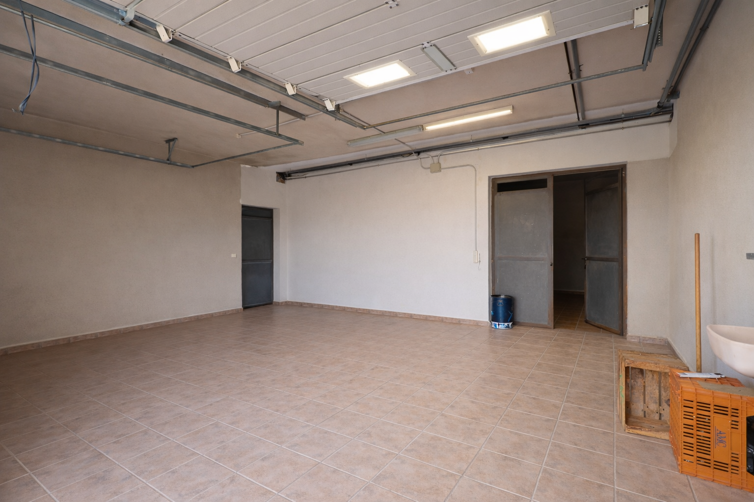 Amplio garaje o sala de servicio con suelo de baldosas claras, techo con luminarias industriales y múltiples accesos. Amplio garaje o sala de servicio con suelo de baldosas claras, techo con luminarias industriales y múltiples accesos.