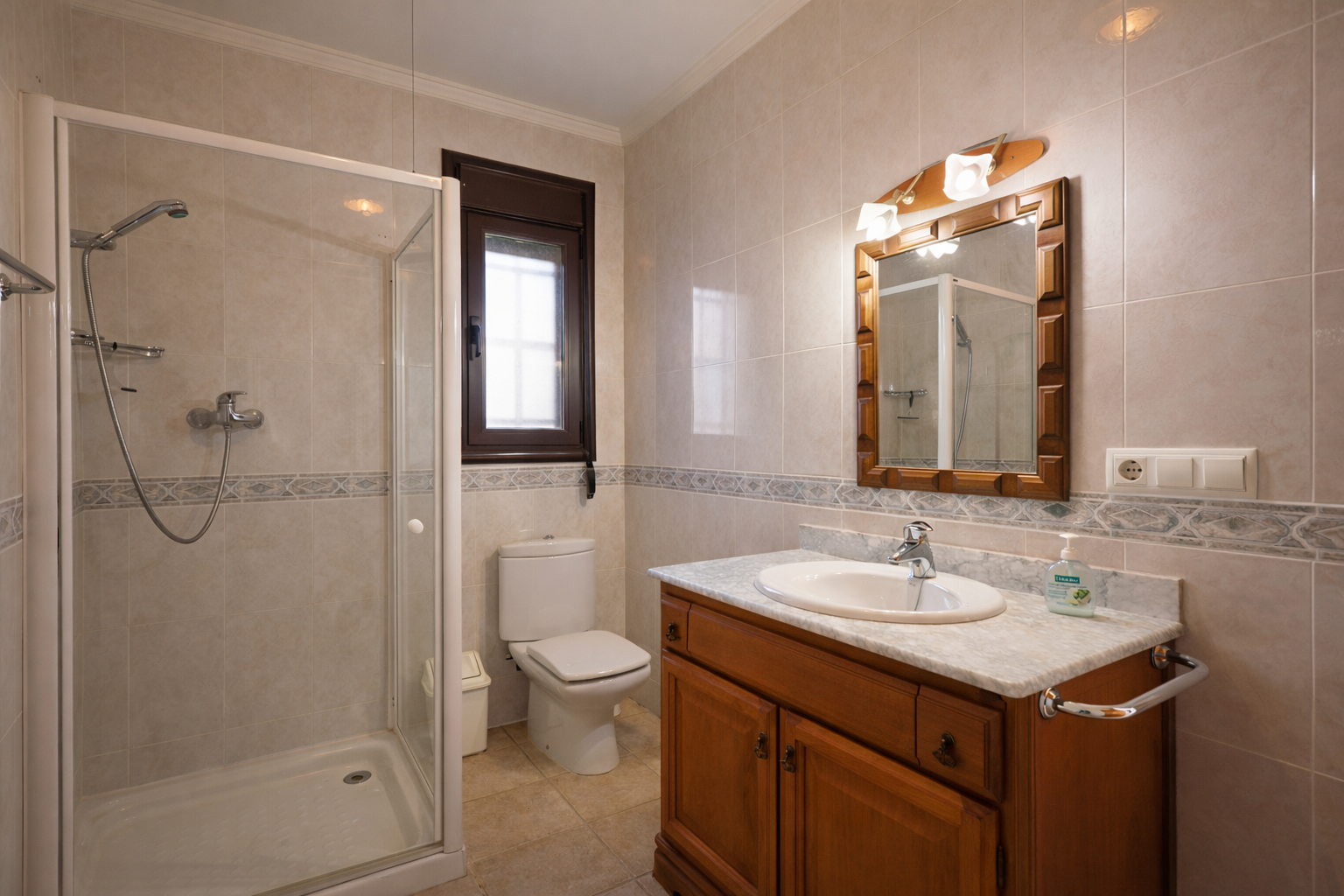 Baño con ducha de mampara, inodoro, y tocador de madera con encimera de mármol. Las paredes están revestidas con azulejos claros y una cenefa decorativa. Baño con ducha de mampara, inodoro, y tocador de madera con encimera de mármol. Las paredes están revestidas con azulejos claros y una cenefa decorativa.