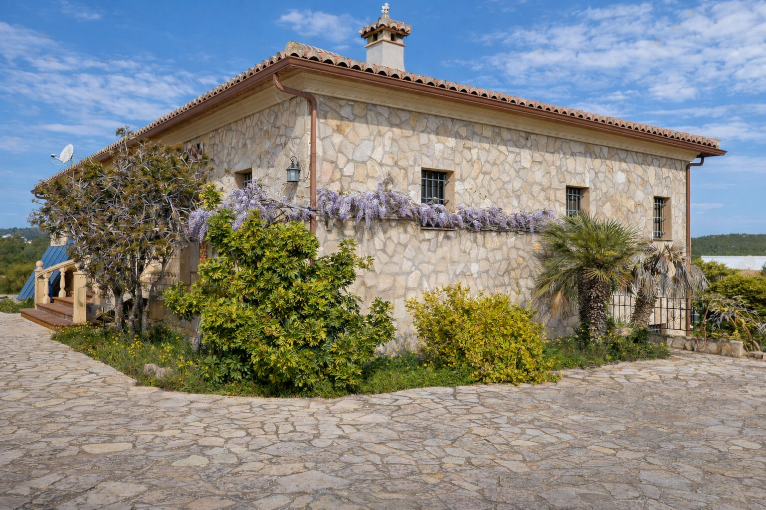 Fachada de piedra rústica con tejado de teja, adornada con glicinias moradas bajo las ventanas enrejadas. El camino de entrada está empedrado. Fachada de piedra rústica con tejado de teja, adornada con glicinias moradas bajo las ventanas enrejadas. El camino de entrada está empedrado.