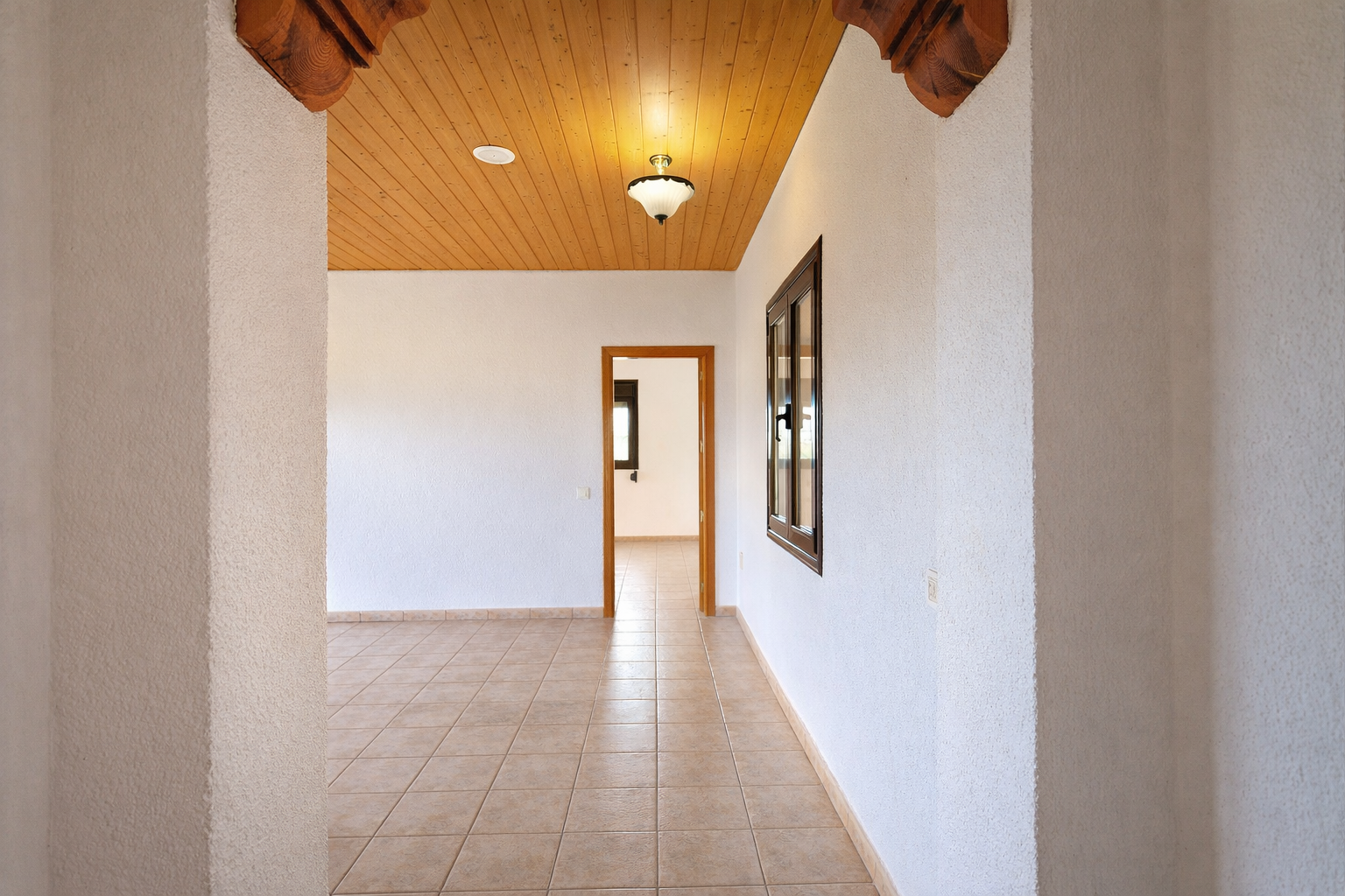 Pasillo interior con suelo de baldosas claras y paredes blancas texturizadas. Techo de madera con iluminación y elementos decorativos tallados en la entrada. Pasillo interior con suelo de baldosas claras y paredes blancas texturizadas. Techo de madera con iluminación y elementos decorativos tallados en la entrada.