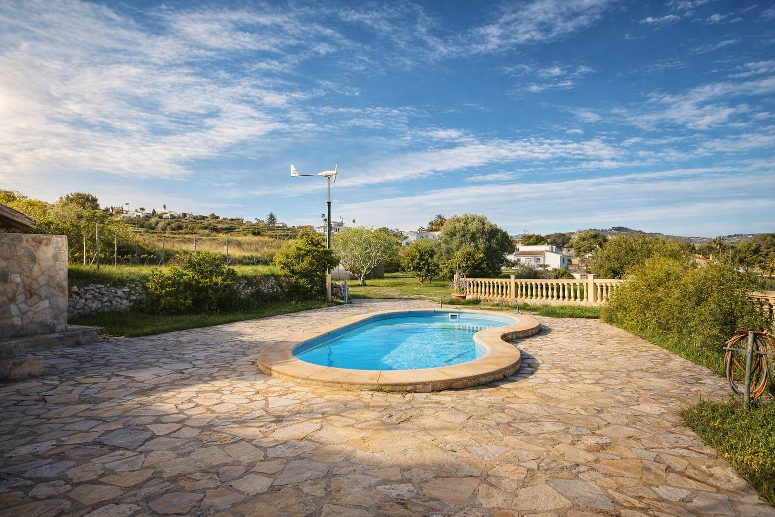 Piscina de forma orgánica rodeada por una amplia terraza de losas irregulares. Al fondo, se aprecian colinas verdes con vegetación y una turbina eólica. Piscina de forma orgánica rodeada por una amplia terraza de losas irregulares. Al fondo, se aprecian colinas verdes con vegetación y una turbina eólica.