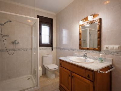Baño con ducha de mampara, inodoro, y tocador de madera con encimera de mármol. Las paredes están revestidas con azulejos claros y una cenefa decorativa.