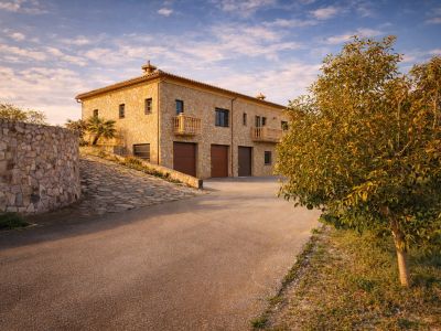 El exterior de la finca, revestido de piedra, presenta tres garajes integrados y balcones elegantes, con acceso por una calzada pavimentada y un muro de piedra curvo.