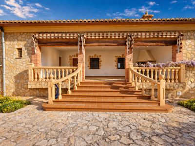 Entrada principal con amplia naya de suelo de terracota, fachada de piedra natural y escalera con balaustrada de estilo clásico. Se aprecia una chimenea y glicinia, además de un panel solar.