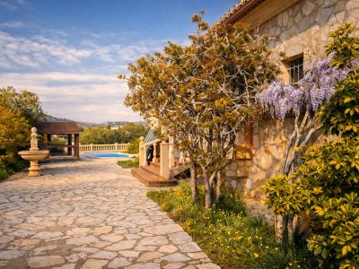 Fachada de piedra de la finca con enredadera de glicinia florecida. Un camino de losas conduce a la piscina y una pérgola de madera, con una fuente de piedra en el jardín.