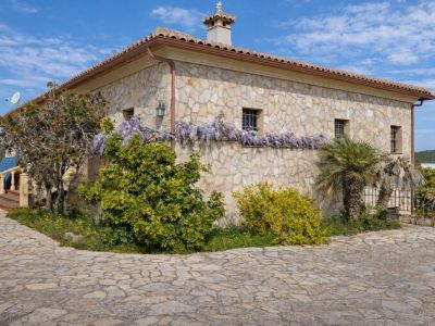 Fachada de piedra rústica con tejado de teja, adornada con glicinias moradas bajo las ventanas enrejadas. El camino de entrada está empedrado.