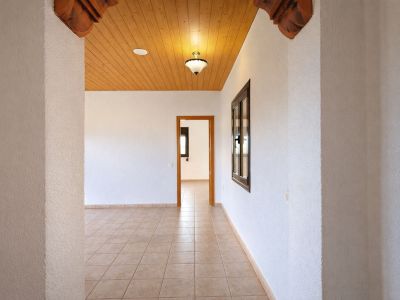 Pasillo interior con suelo de baldosas claras y paredes blancas texturizadas. Techo de madera con iluminación y elementos decorativos tallados en la entrada.