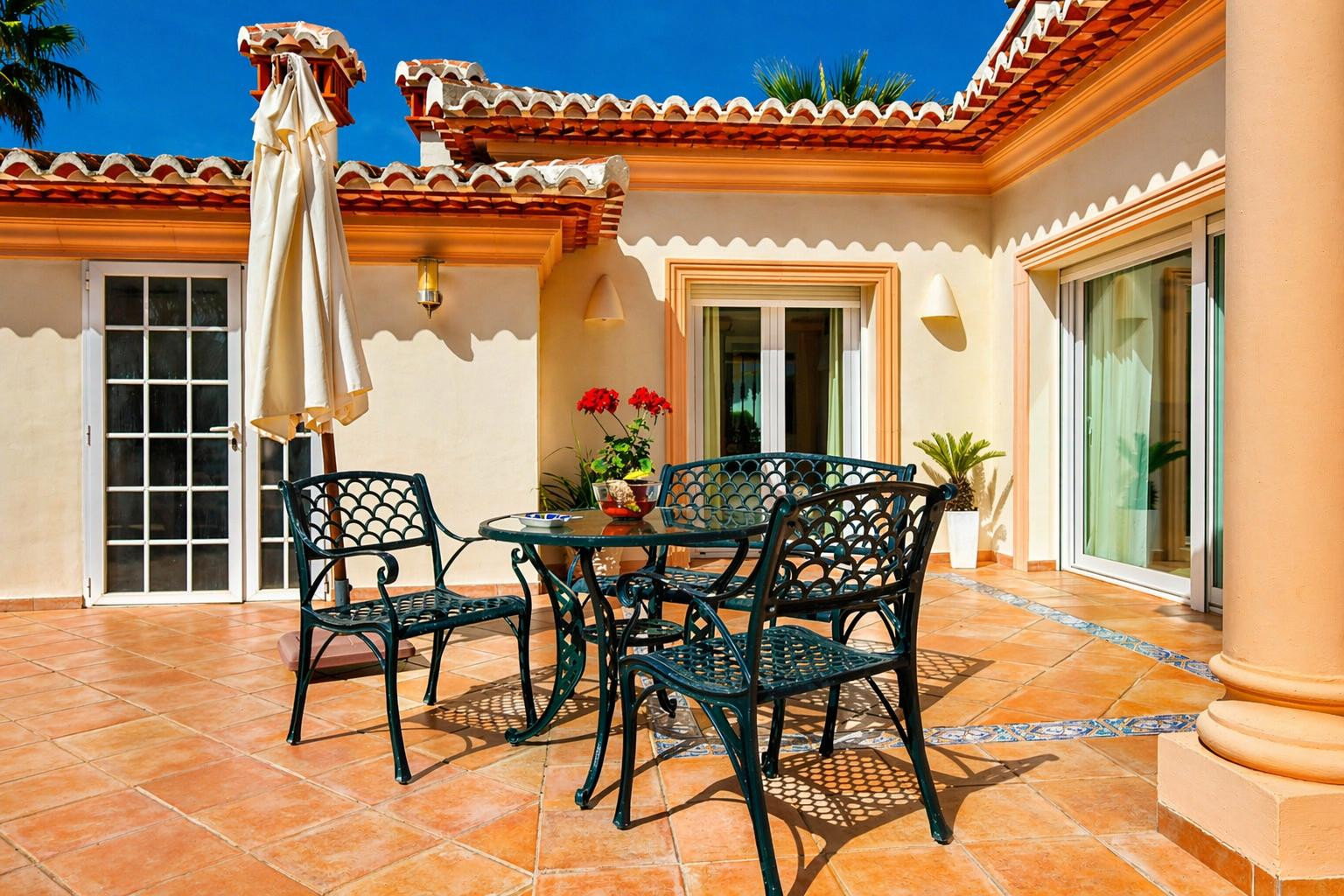 Amplia terraza exterior con suelo de terracota y un borde de azulejos decorativos. Destaca un conjunto de comedor de metal verde oscuro y elementos arquitectónicos mediterráneos.