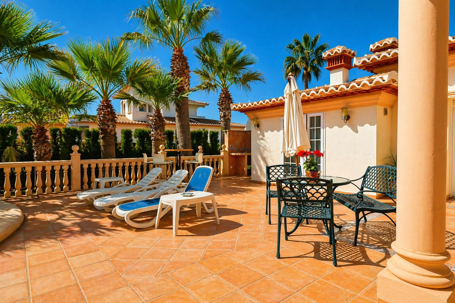 Amplia terraza exterior pavimentada con baldosas de terracota, equipada con tumbonas y un conjunto de mesa y sillas de metal bajo una sombrilla. Palmeras y una balaustrada de piedra enmarcan el espacio.