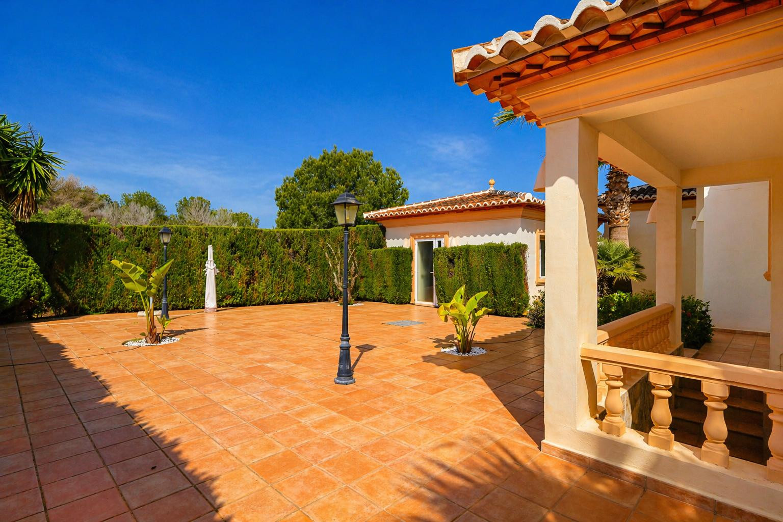 Amplio patio exterior con baldosas de terracota, delimitado por un seto verde, que incluye farolas clásicas y un porche cubierto con balaustrada y tejado tradicional.