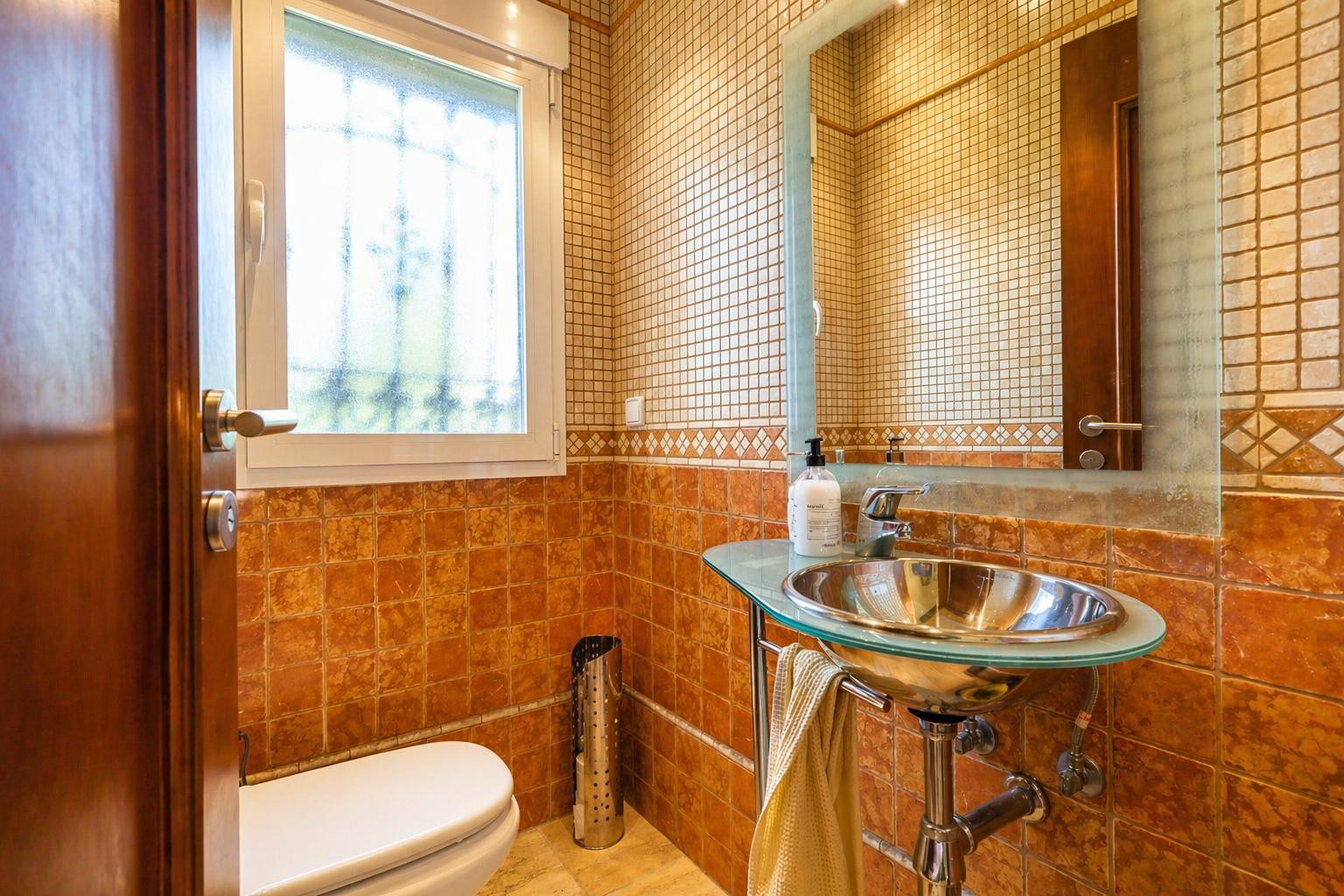 Aseo de cortesía con azulejos terracota y mosaico, lavabo moderno con encimera de cristal y bol metálico, y ventana que aporta luz natural.