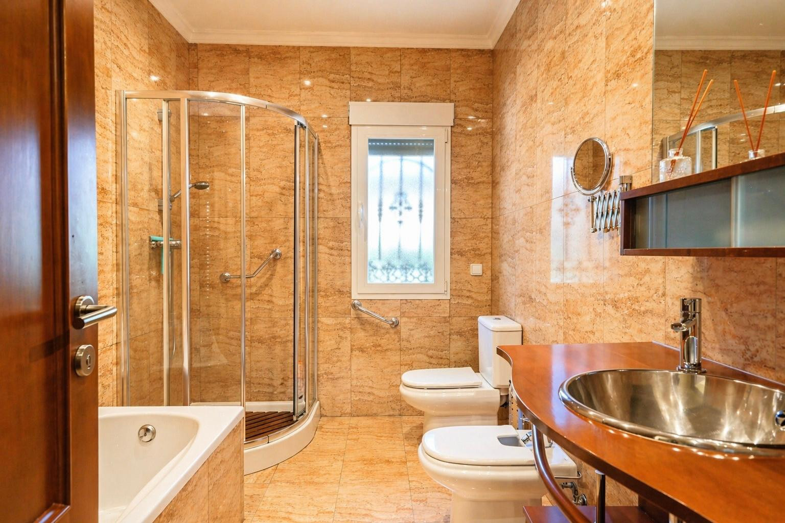 Baño completo con azulejos de tonos tierra, bañera, ducha de cristal curvada con barras de apoyo, inodoro, bidé y lavabo de acero inoxidable sobre mueble de madera.