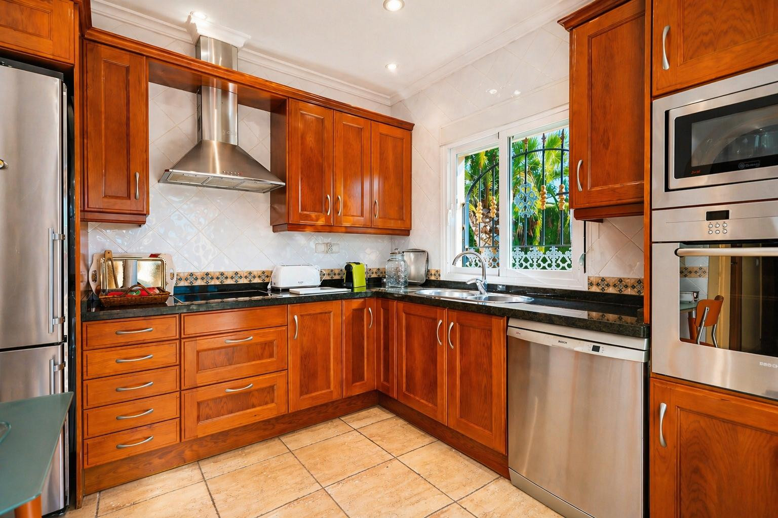 Cocina funcional con armarios de madera cálida, encimeras de granito oscuro y electrodomésticos de acero inoxidable integrados. Ventana con reja decorativa y vistas al exterior.