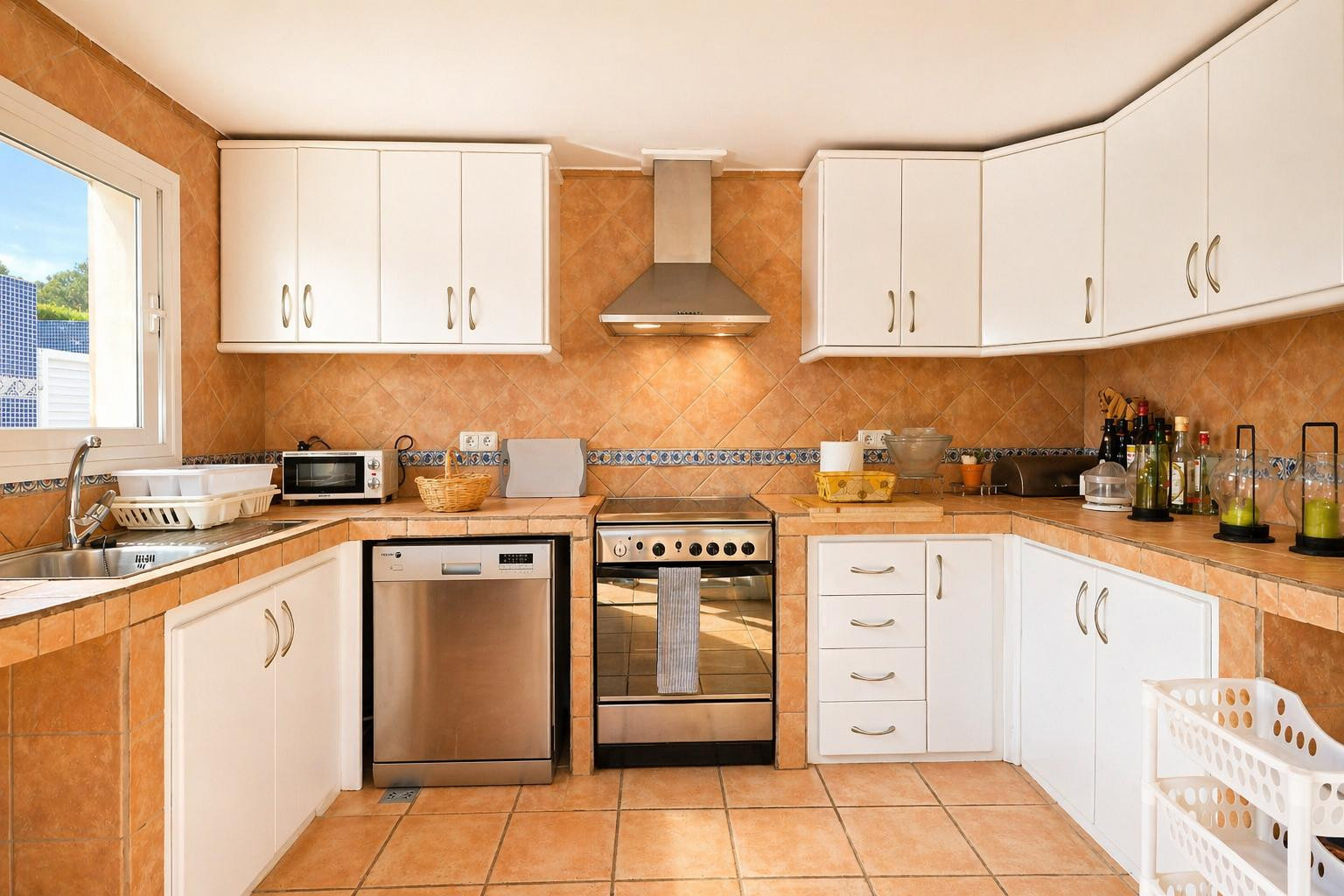 Cocina funcional equipada con azulejos de terracota en suelo y pared, armarios blancos y electrodomésticos de acero inoxidable. La luz natural entra por la ventana sobre el fregadero.